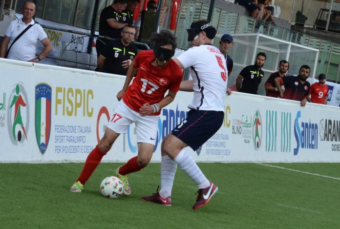 Two male blind football players in action. One athlete is wearing a red jersey and dribbles the ball past an opponent.